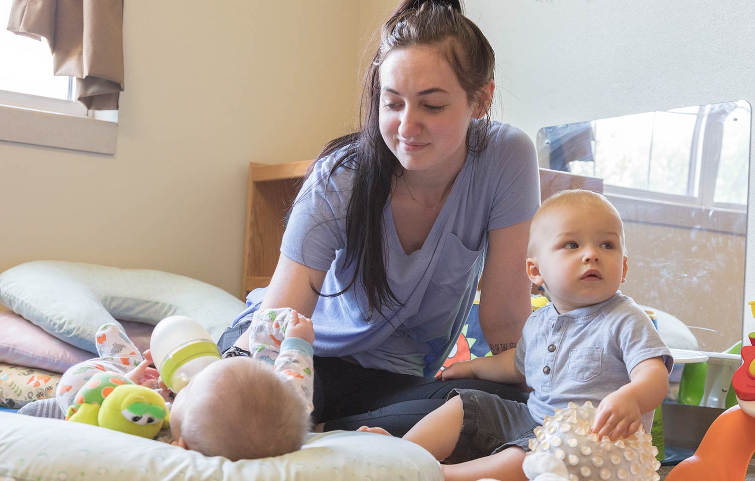 A teacher sitting on the ground caring for an infant and a toddler